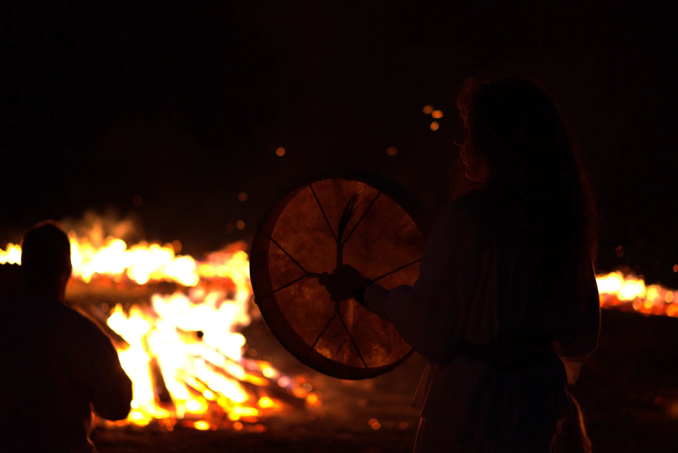 A drummer holding a frame drum beside the ceremonial fire